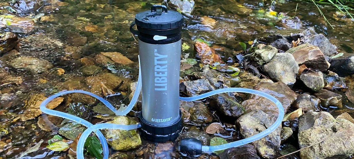 Blue LifeSaver Liberty Purifier with drinking tube coiling around it, in a shallow stream on wet rocks