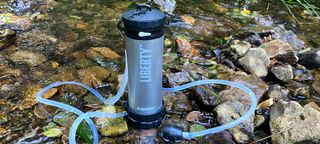 Blue LifeSaver Liberty Purifier with scavenger hose coiling around it, in a shallow stream on wet rocks