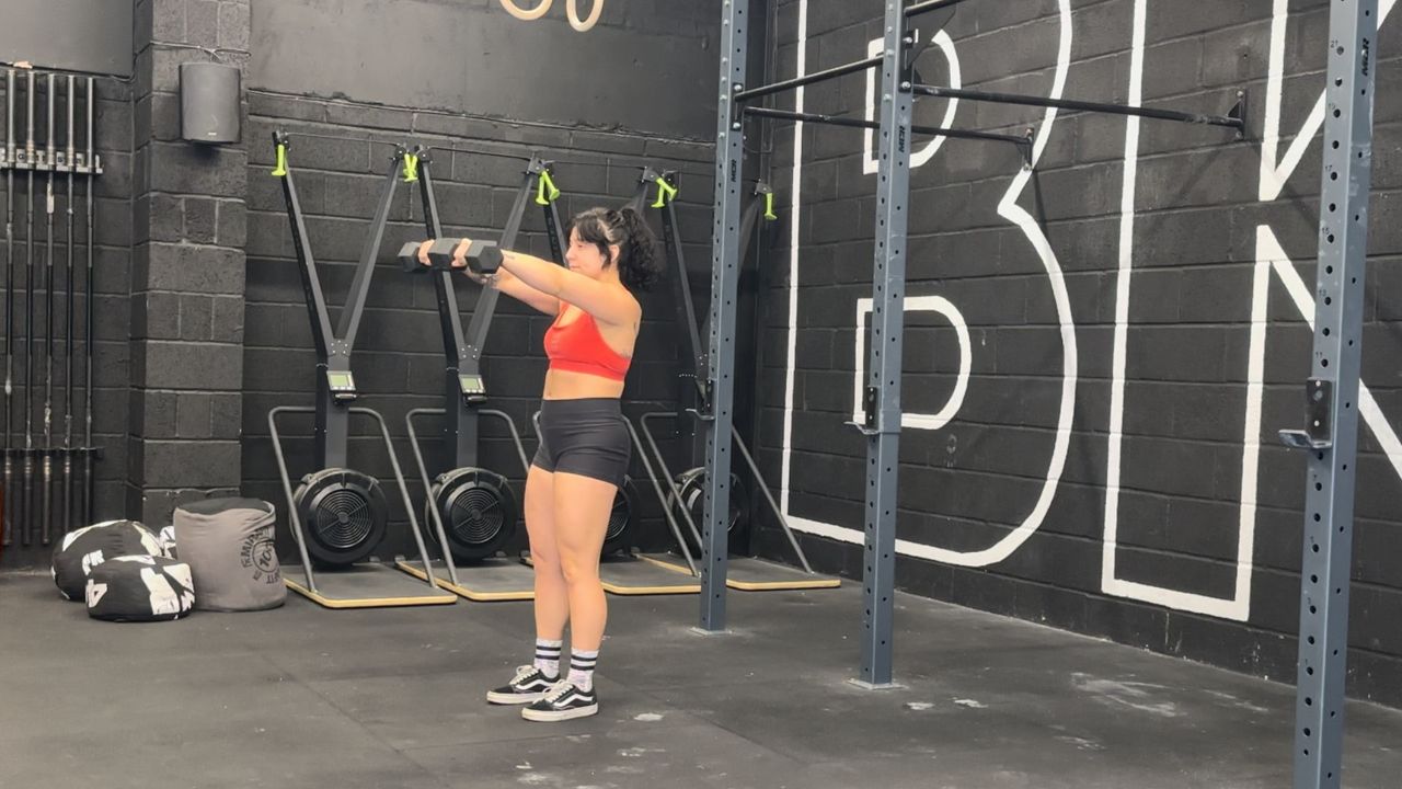 Raquel Sanjurjo performs a lateral to front raise. She is standing in a gym holding a pair of dumbbells out the sides, with her arms straight in front.
