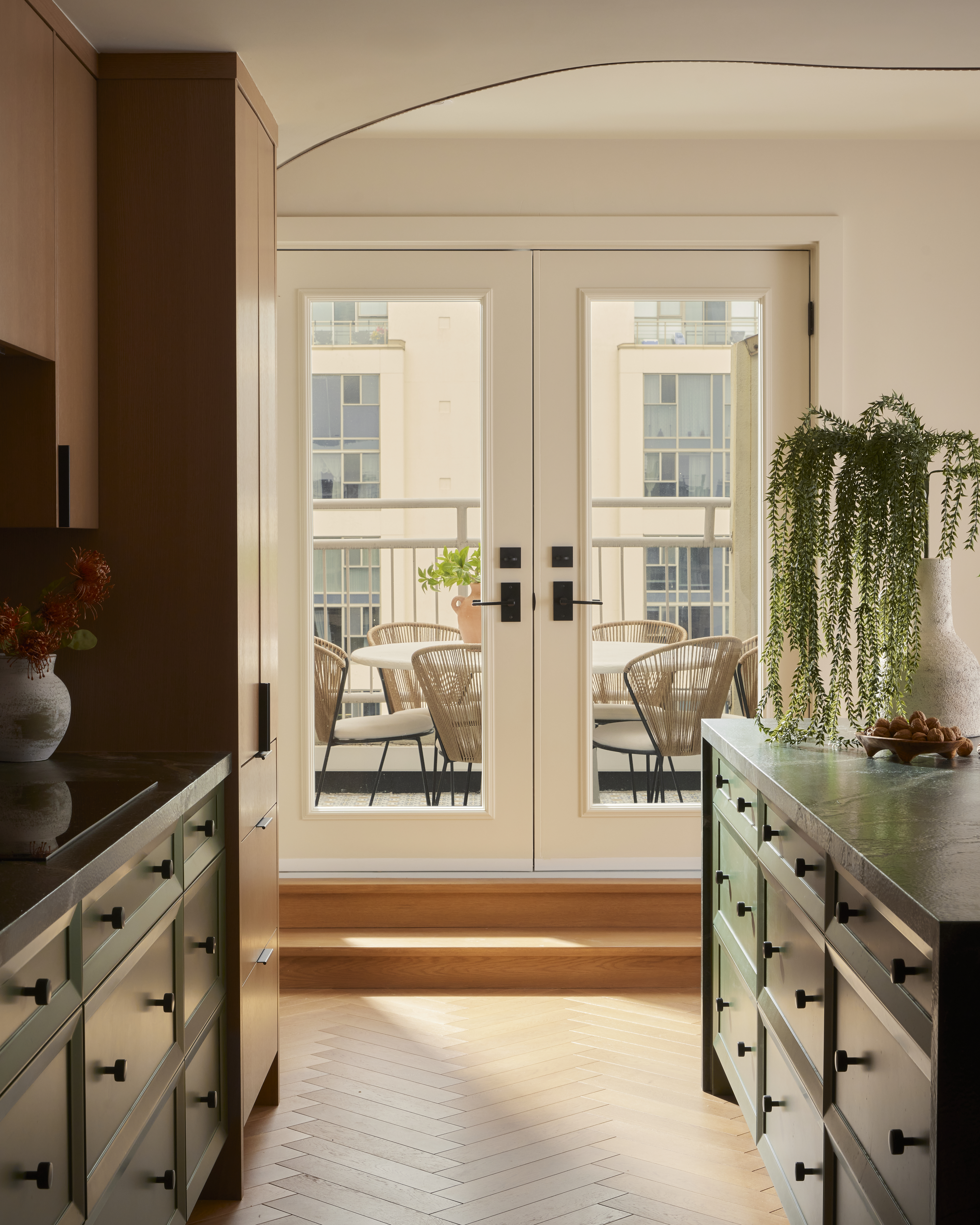 A green-painted kitchen with a kitchen island leads onto a minimalist terrace with rattan and beige fabric chairs and a white table, and catches sunshine on its herringbone terracotta floors.