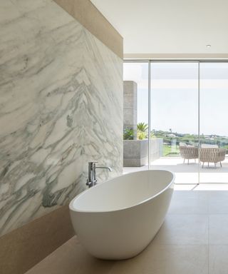 modern bathroom with one marble wall, modern tub, view of outside, stone flooring,