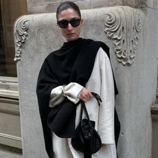 andrea wears white coat and black cape scarf while holding black handbag and posing with sunglasses