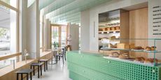 A minimalist coffee shop and bakery boasts a chequered, resin bar counter in a minty shade of green complete with freshly baked pastries and bread kept in a glassy display, chrome accents, and a Scandinavian, wood-clad atmosphere.