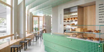 A minimalist coffee shop and bakery boasts a chequered, resin bar counter in a minty shade of green complete with freshly baked pastries and bread kept in a glassy display, chrome accents, and a Scandinavian, wood-clad atmosphere.