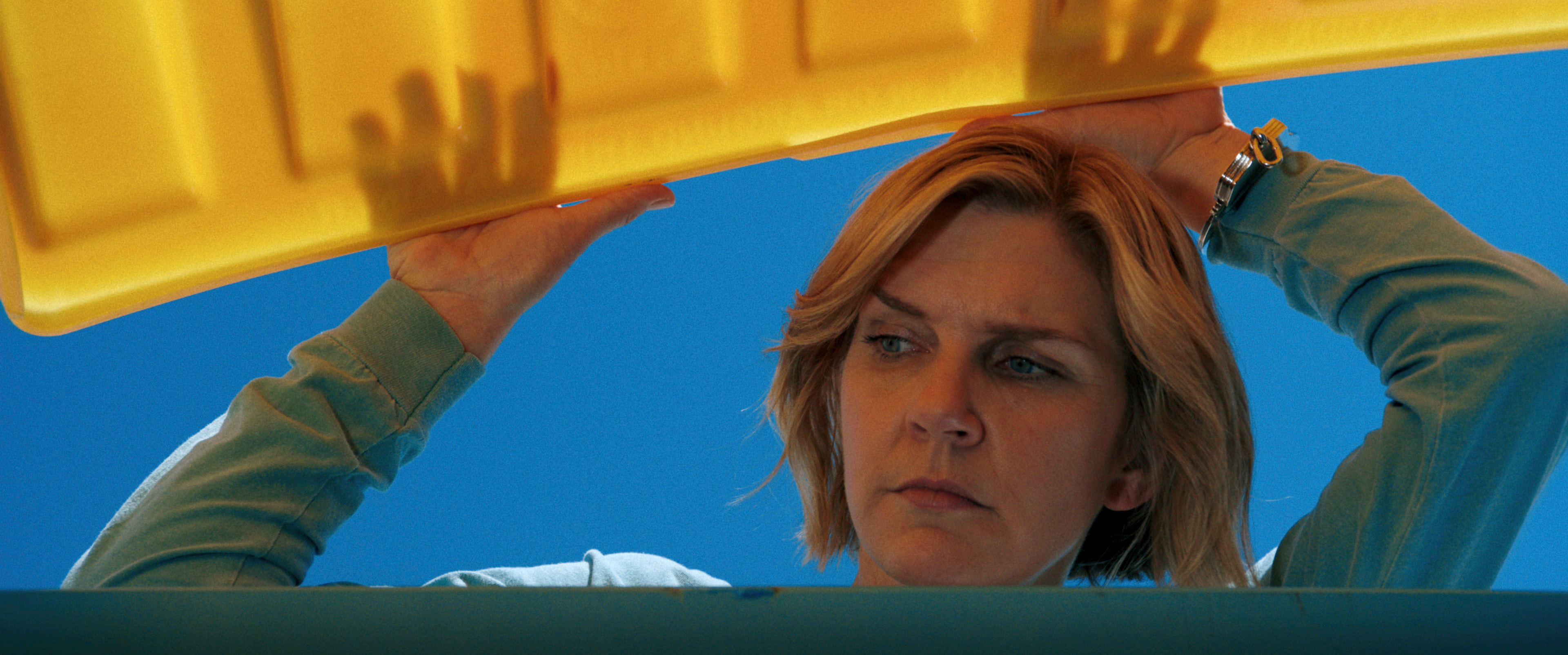 A blond woman (Rhea Seehorn as Carol) lifts a yellow trashbin lid and peeks inside as the blue sky shines behind her, in a still from 'Pluribus' season 1.