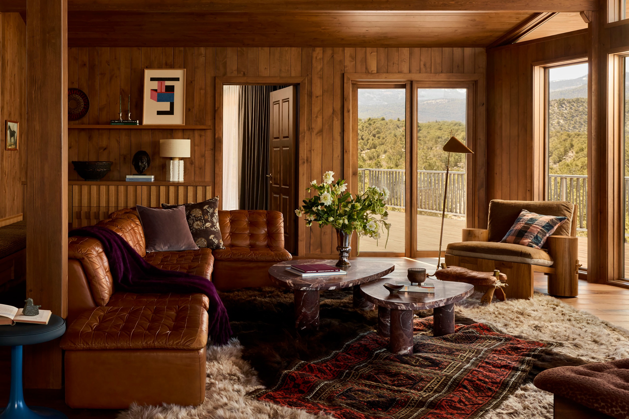 A warm, modern rustic living room clad in wood paneling, layered with rugs, textures, throw blankets and cushions on a brown leather sofa with a modern armchair in the corner by the windows