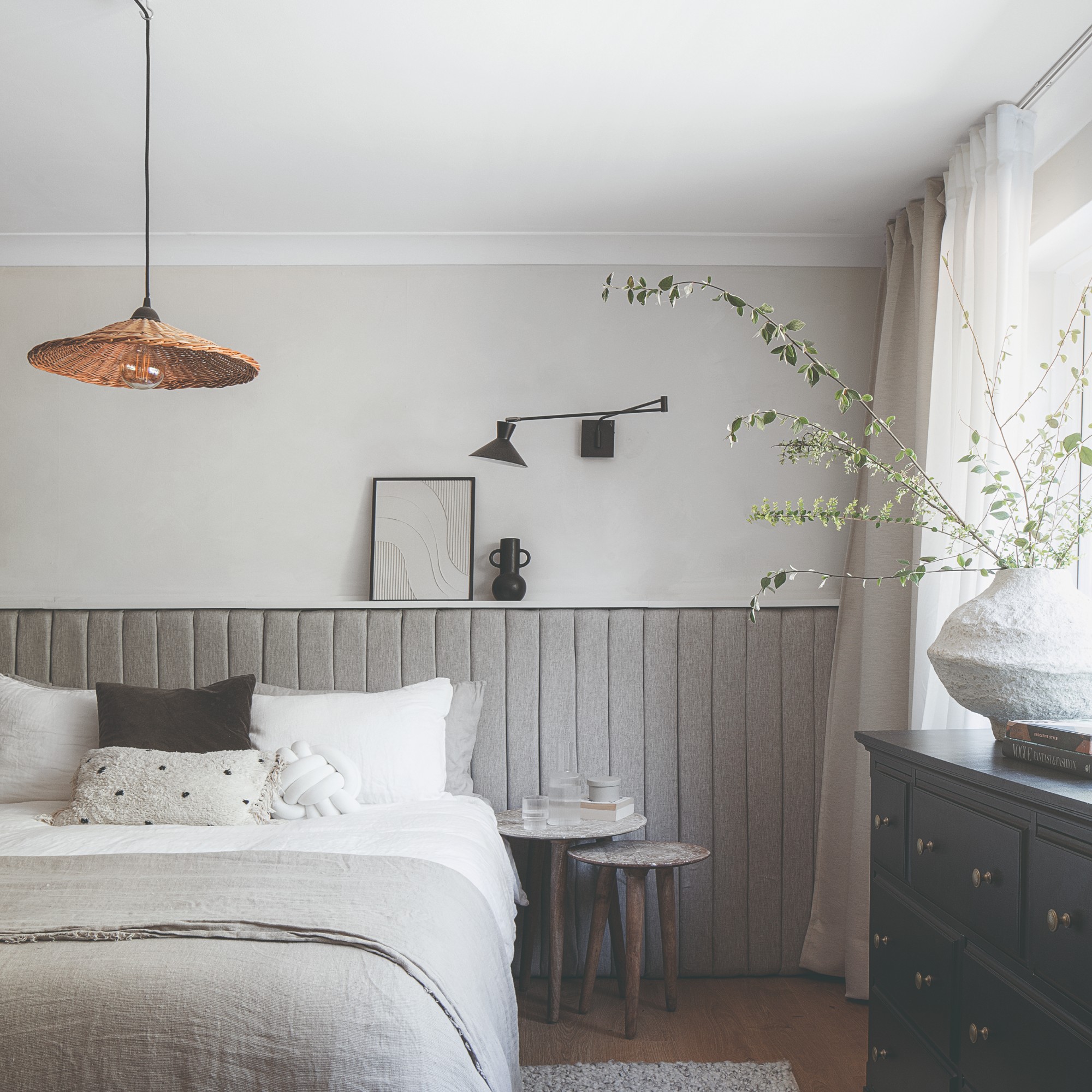 A neutral living room with half of the wall covered in a quilted padding in grey, with a black adjustable wall light and a rattan pendant light