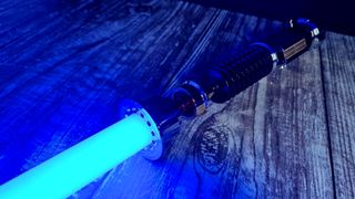 A lit SabersPro Obi Wan EP3 blade, shining blue on a wooden surface