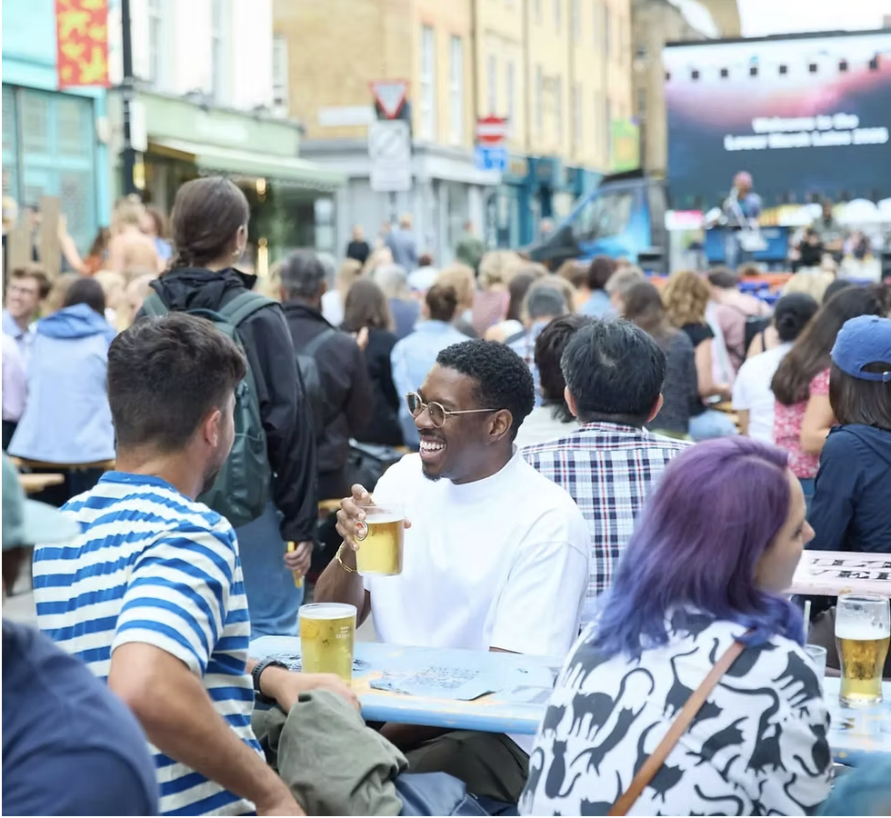 an image of people gathered outside in the outdoor market sipping on drinks on tables as live music plays behind them