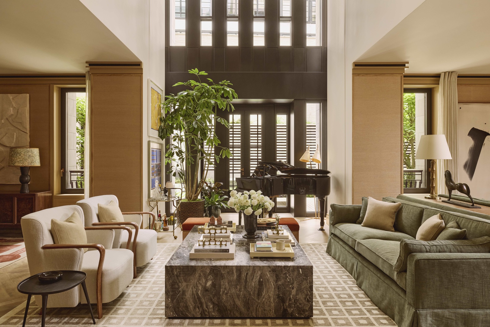 a modern living room with green sofa, marble coffee table styled with books, cheese, vase of flowers, and two cream armchairs on the opposite side, with a side table, large geometric-print rug, shutters on windows, double height ceiling, potted tree and piano
