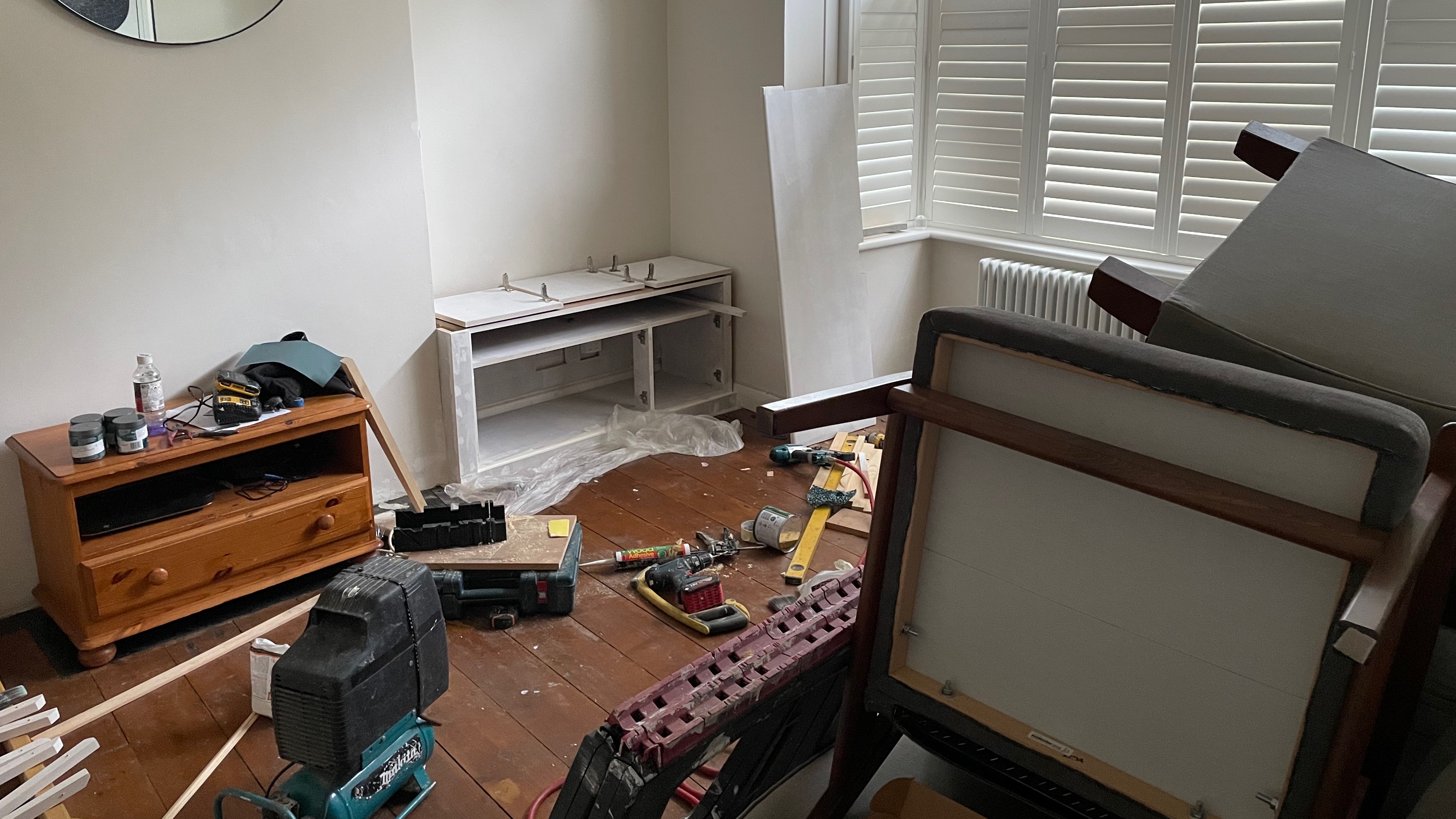A front room in all sorts of disarray, with tools strewn over the floor, chairs place on a sofa to keep them out of the way, and so on.