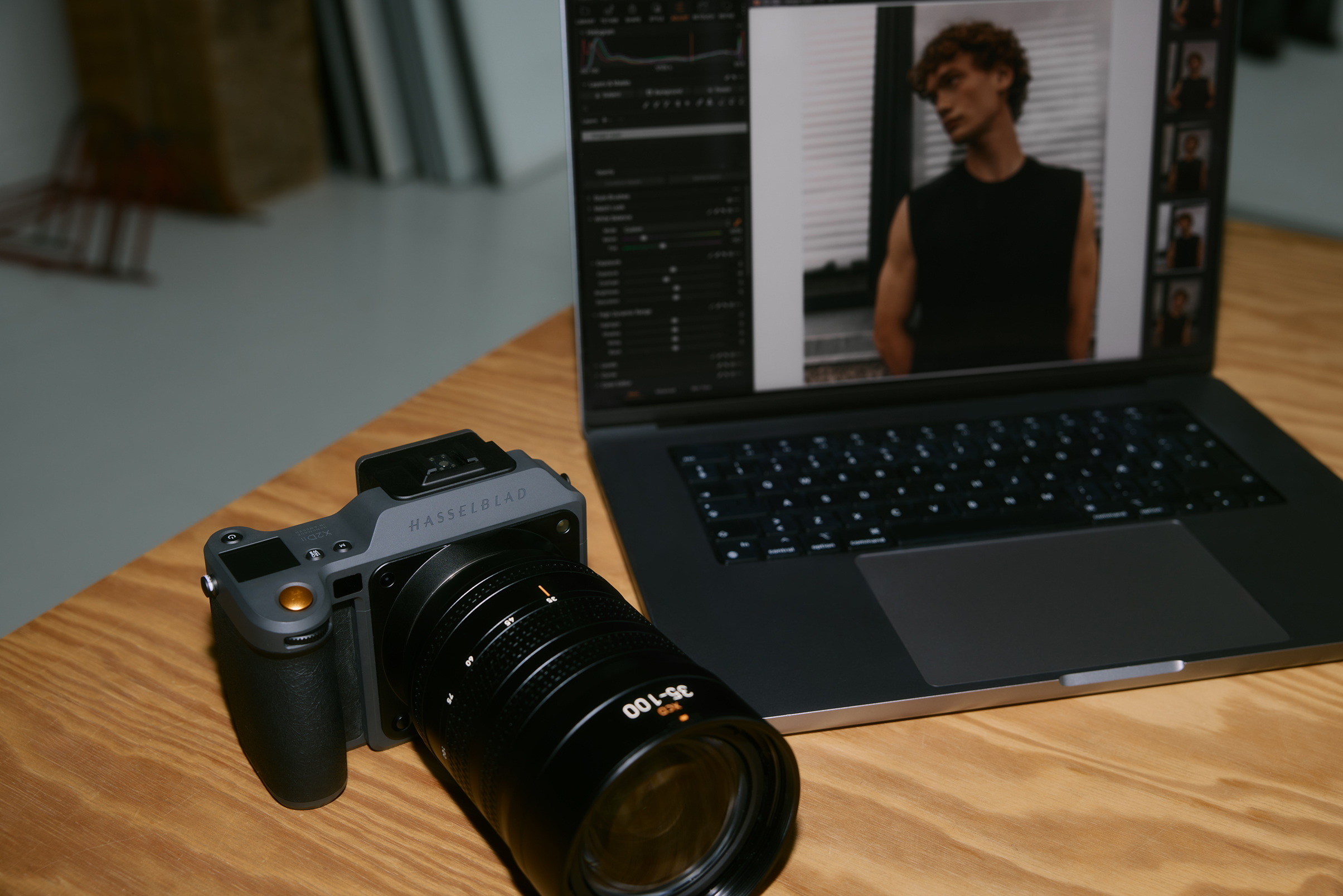 Camera with large lens on a wooden table next to a laptop displaying a portrait photo of a man in editing software