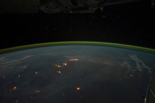 Wildfires and perhaps some intentionally set agricultural fires burn on the continent of Australia, with smoke plumes faintly visible in the night sky. A gold and green halo of atmospheric airglow hangs above the horizon in the distance.
