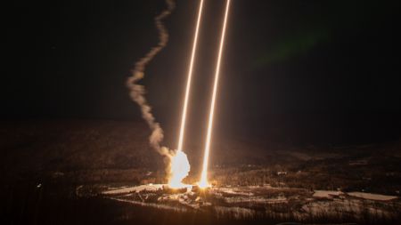 streaks of bright light reach from the ground to the sky above a snowy landscape dotted with evergreen trees