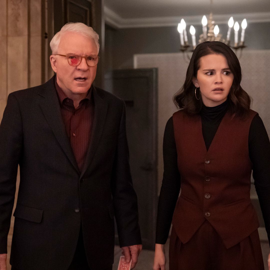 A man (Steve Martin as Charles) wearing glasses with red marker on the left lens stands with a woman (Selena Gomez as Mabel) in a ballroom doorway with a chandelier behind them, in &#039;Only Murders in the Building.&#039;