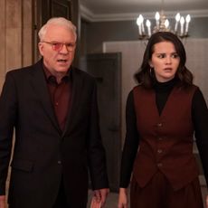 A man (Steve Martin as Charles) wearing glasses with red marker on the left lens stands with a woman (Selena Gomez as Mabel) in a ballroom doorway with a chandelier behind them, in 'Only Murders in the Building.'