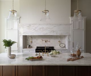 A white kitchen with marble countertops and range hood and a wooden kitchen island. Throughout the kitchen, unlacquered brass hardware has been used for a timeless look