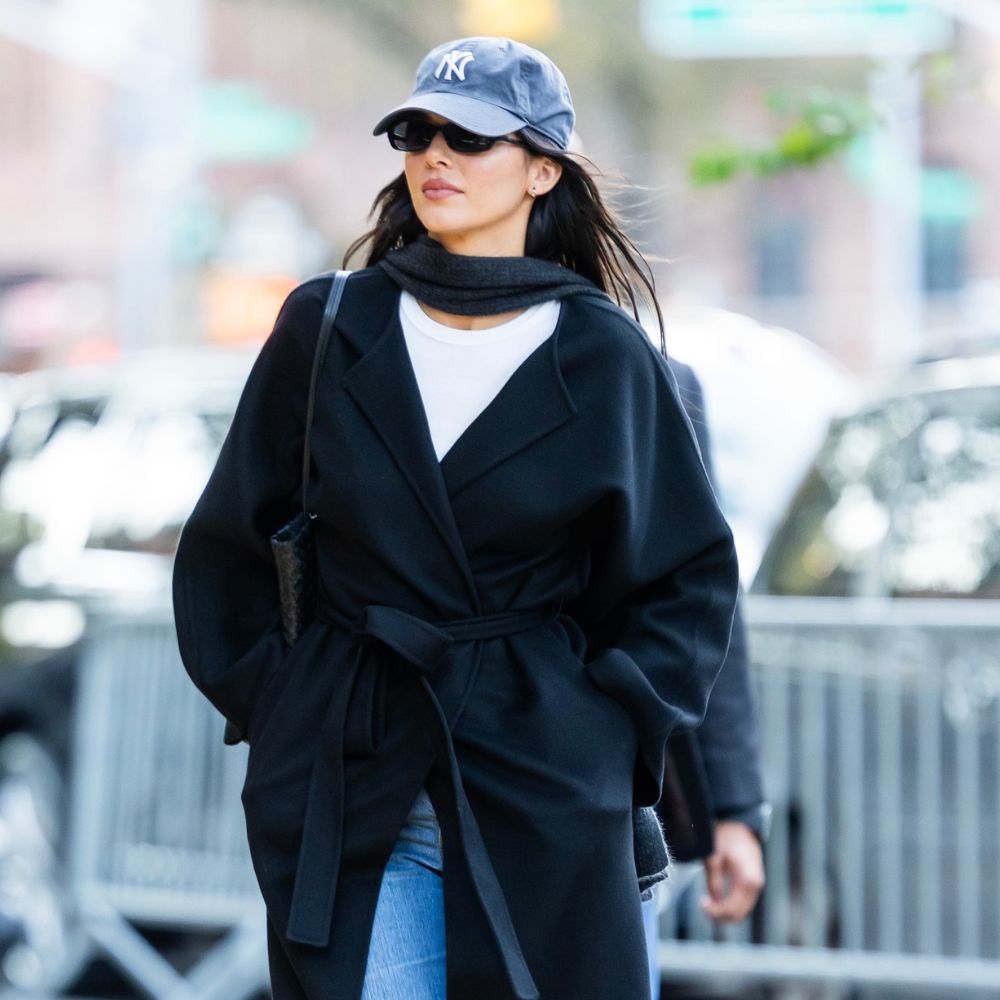 Kendall Jenner walks down the street wearing bootcut jeans with pointed-toe boots and a black belted coat, grey scarf and cap.