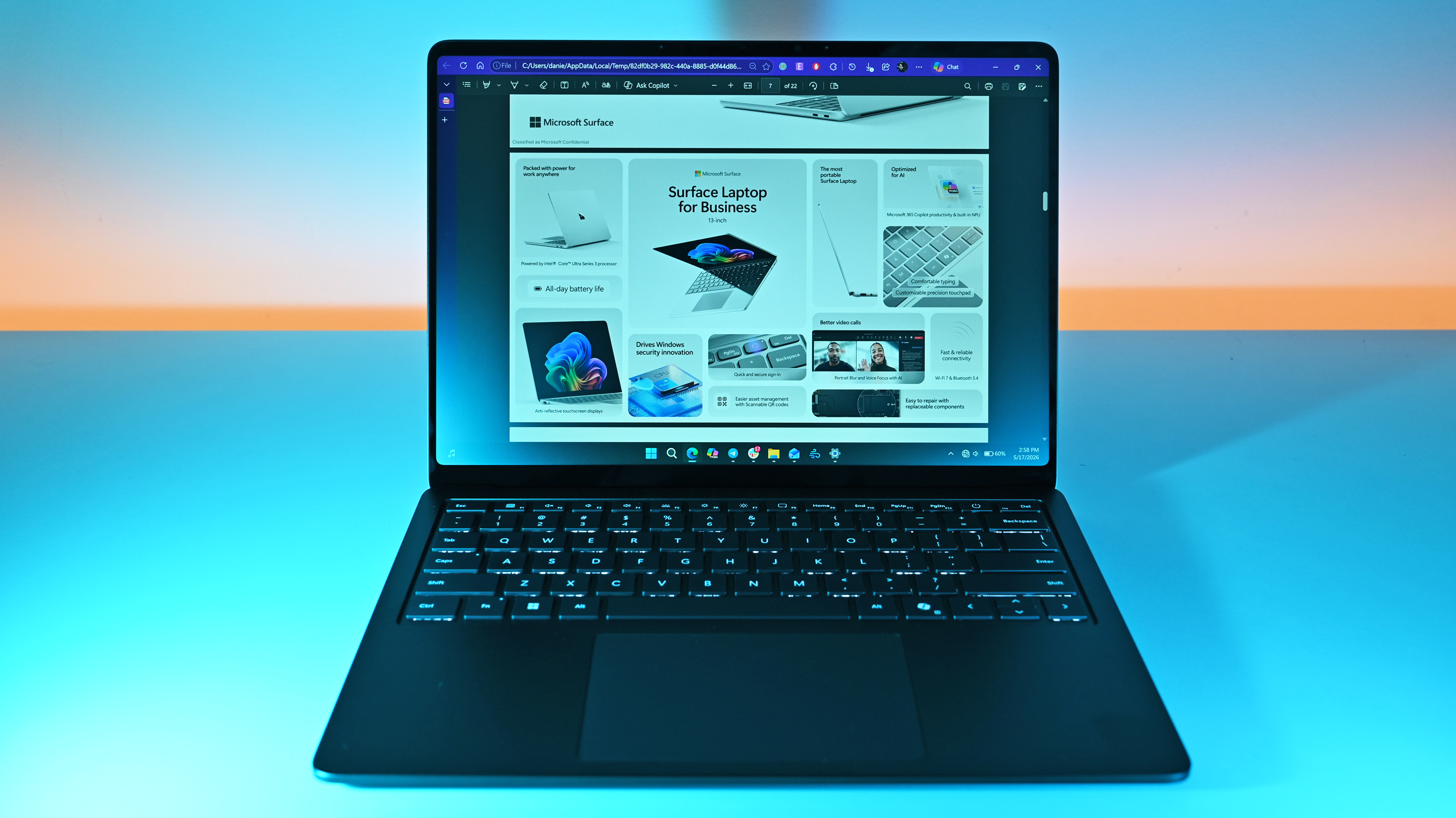 Laptop on a desk displaying a webpage about Surface Laptop for Business. The screen shows colorful product images and text. Soft pink and blue lighting in the background.