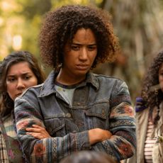 jasmine savoy brown crosses her arms while looking at something in the wilderness in yellowjackets season 3 