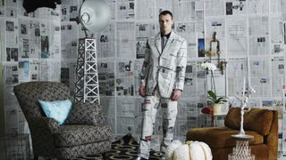 A man dressed in a newspaper suit stands in front of a newspaper-covered wall.