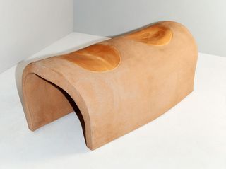 View of the ‘Johnny Guitar’ bench - an arch shaped terracotta coloured bench pictured in a room with a grey floor and walls