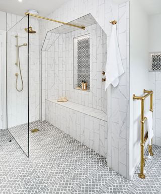 Tiled bathroom with gray patterned floor tiles, marble tiles in brick design on walls, large shower with glass screen, seating area, gold fixtures and fittings