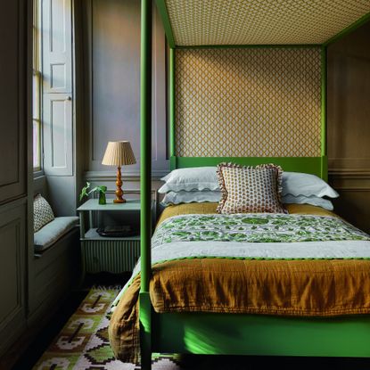 A light brown-painted bedroom with a green poster bed and a striped mustard yellow table lamp on the bedside table