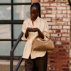 Woman in beige button-down with brown pleated trousers and a suede bag on her phone. 