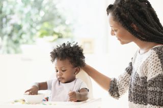 Woman watching baby daughter eat cereal in high chair