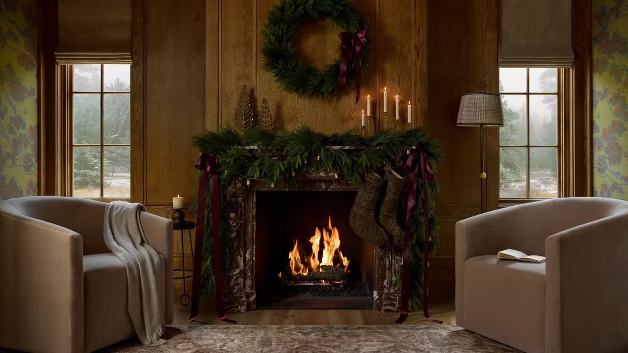 Cozy, festive living room featuring a roaring fire styled with garland and a Christmas wreath overhead. Two sitting chairs flank the wood-paneled scene.