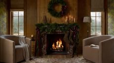 Cozy, festive living room featuring a roaring fire styled with garland and a Christmas wreath overhead. Two sitting chairs flank the wood-paneled scene.