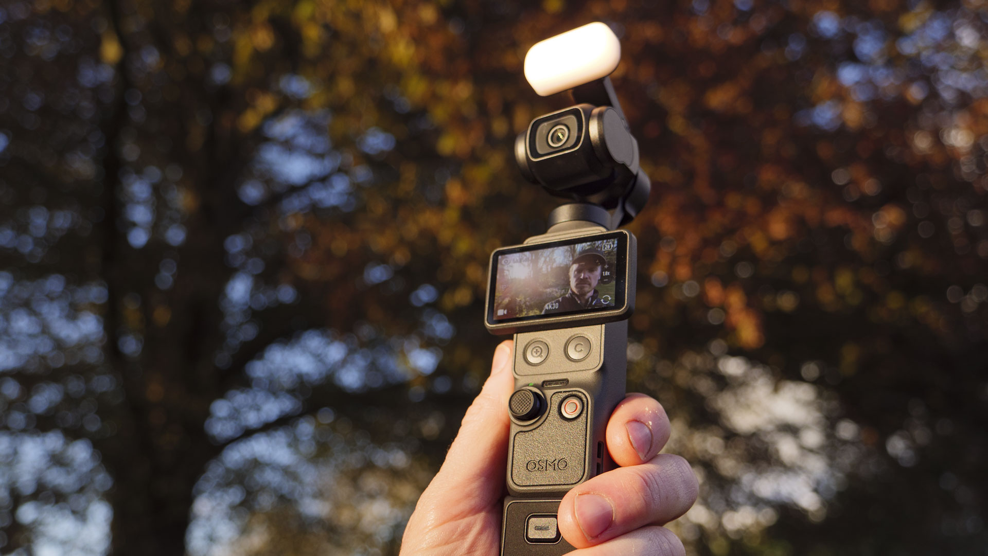 DJI Osmo Pocket 4 vlogging camera with natural background at golden hour, and the user in the frame of the screen