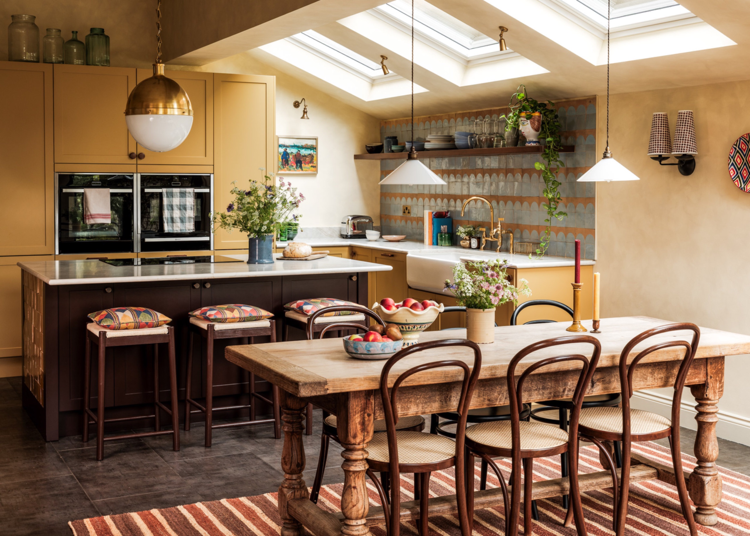 Kitchen with mustard-yellow cabinets and a dark island with cushioned stools, positioned parallel to a wooden dining table with bentwood chairs beneath skylights.