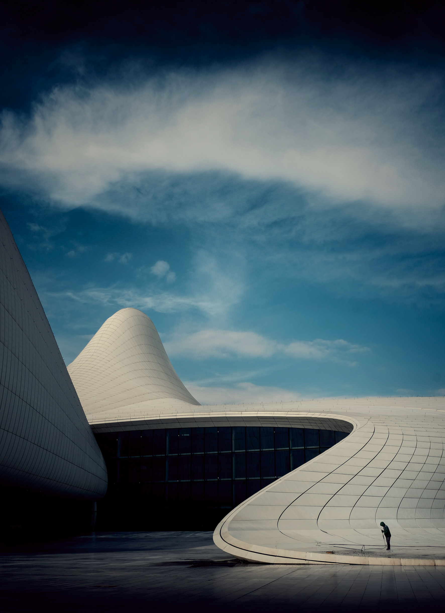 Under a deep blue sky streaked with cloud, the flowing white curves of a futuristic building rise like waves, while a tiny hooded figure holding a hose stands at the base of one sweeping wall.
