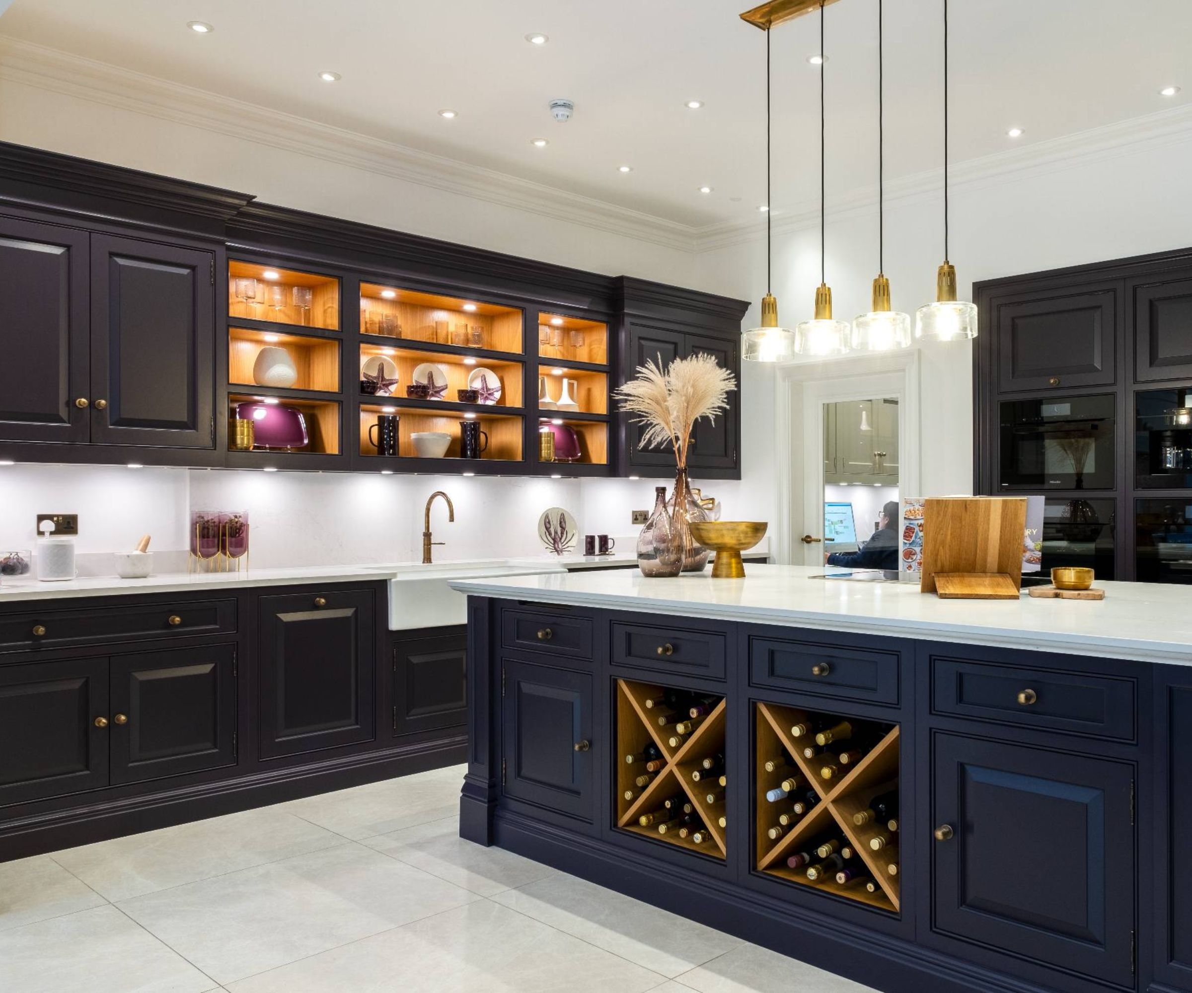 A sophisticated kitchen with deep navy in-frame cabinets and a large central island. Glass-fronted cupboards glow with warm lighting. Brass handles, pendant lights and a built-in wine rack add elegance to the contemporary, high-end design.