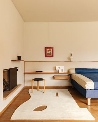 An off-white bedroom with a blue and white bed that has a wooden and white headboard that extends to the wall. There is a white cutout rug next to the bed and a fireplace that is built into the wall on the right side of the bed.