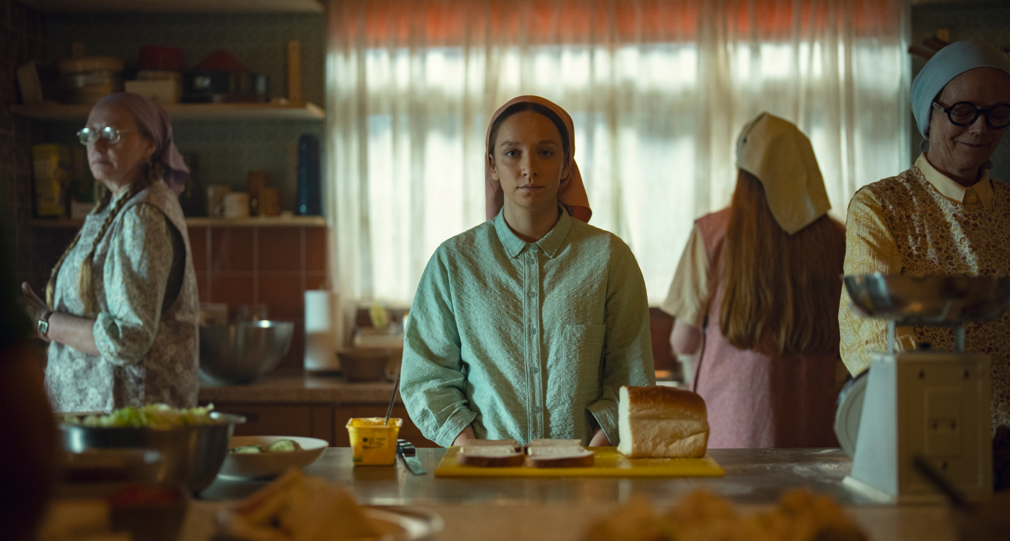 a woman in traditionalist clothing sits at a table slicing bread in a still from netflix's unchosen
