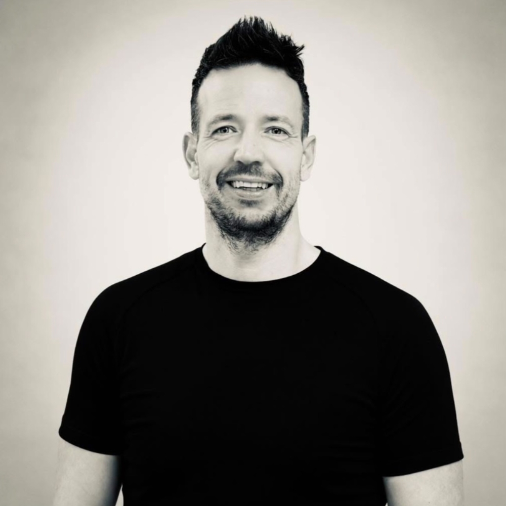 Black-and-white photo of a smiling man with spiked hair and a beard, wearing a black t-shirt.