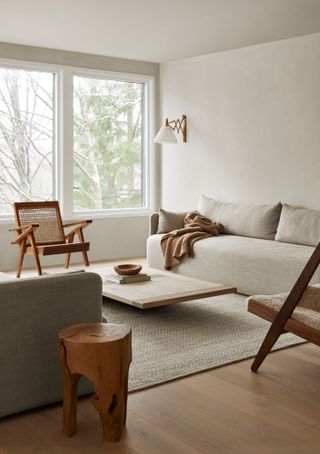 a minimalist living room with a white sofa with beige throw, a low timber coffee table, a rattan armchair under the window, a stump side table and beige rug