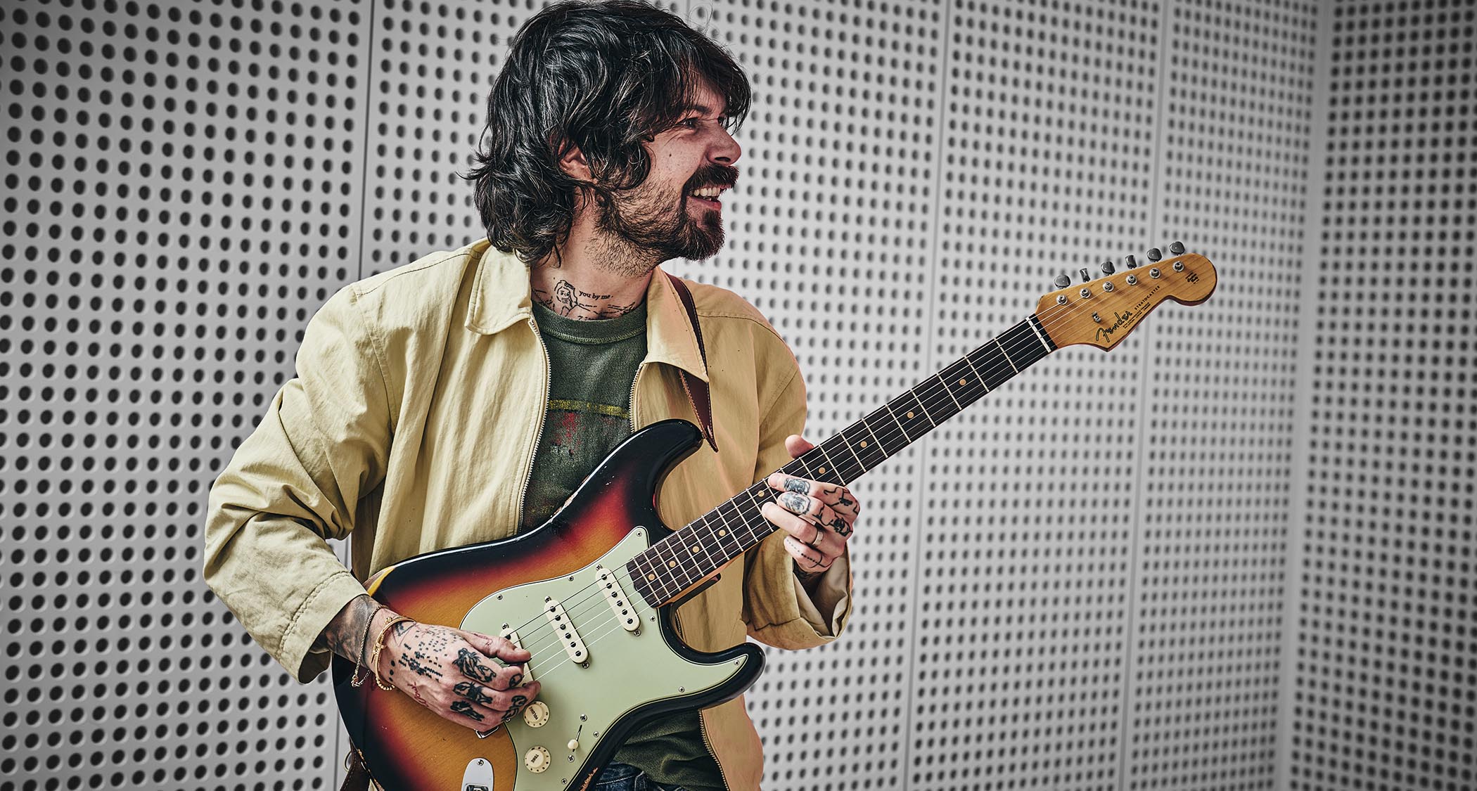 Simon Neil of Biffy Clyro wears a custard-colored jacket and green T-shirt and poses with a sunburst Fender Stratocaster