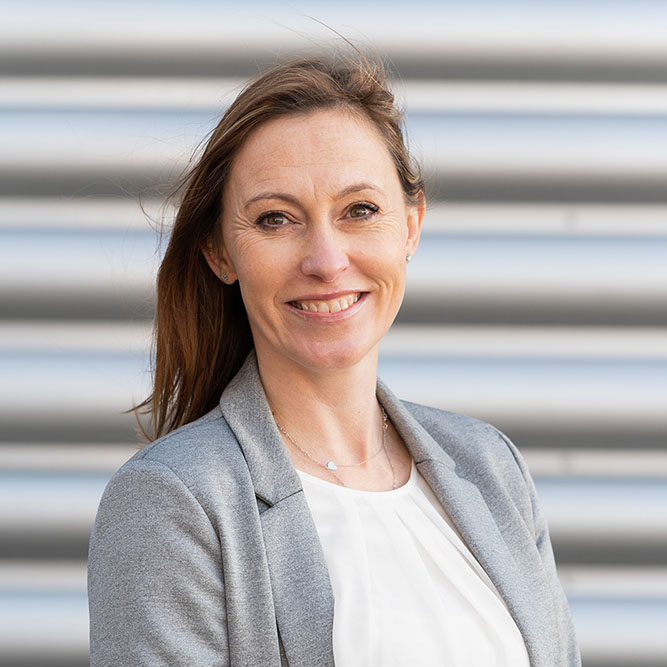 female with shoulder length brown hair stood against grey metal background wearing grey jacket and cream top