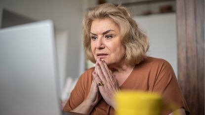 An older woman looks concerned as she looks at her laptop while sitting at her kitchen table.