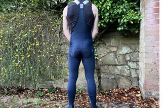 The Rapha Pro Training Tights are being worn by a white male in a sleeveless undervest, he is standing infront of a stone wall with greenery overhanging