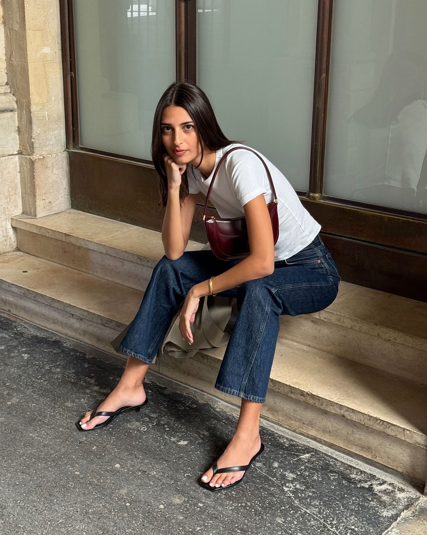 Influencer sits on steps outside wearing a white t-shirt with dark denim flared jeans and heeled flip-flops.