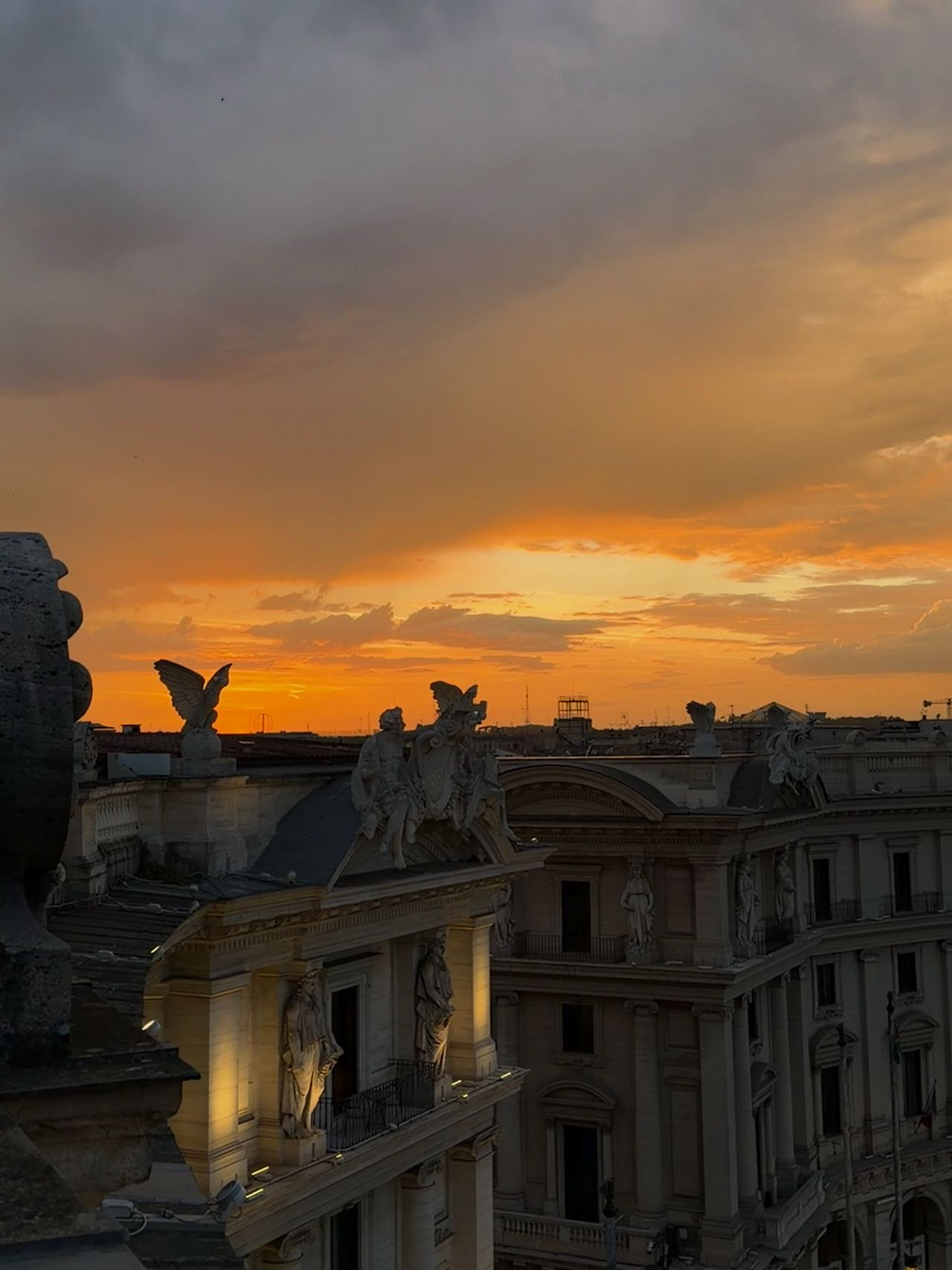 Sunset over Rome viewed from a rooftop terrace, with golden skies glowing behind ornate statues and historic buildings illuminated in the evening light.