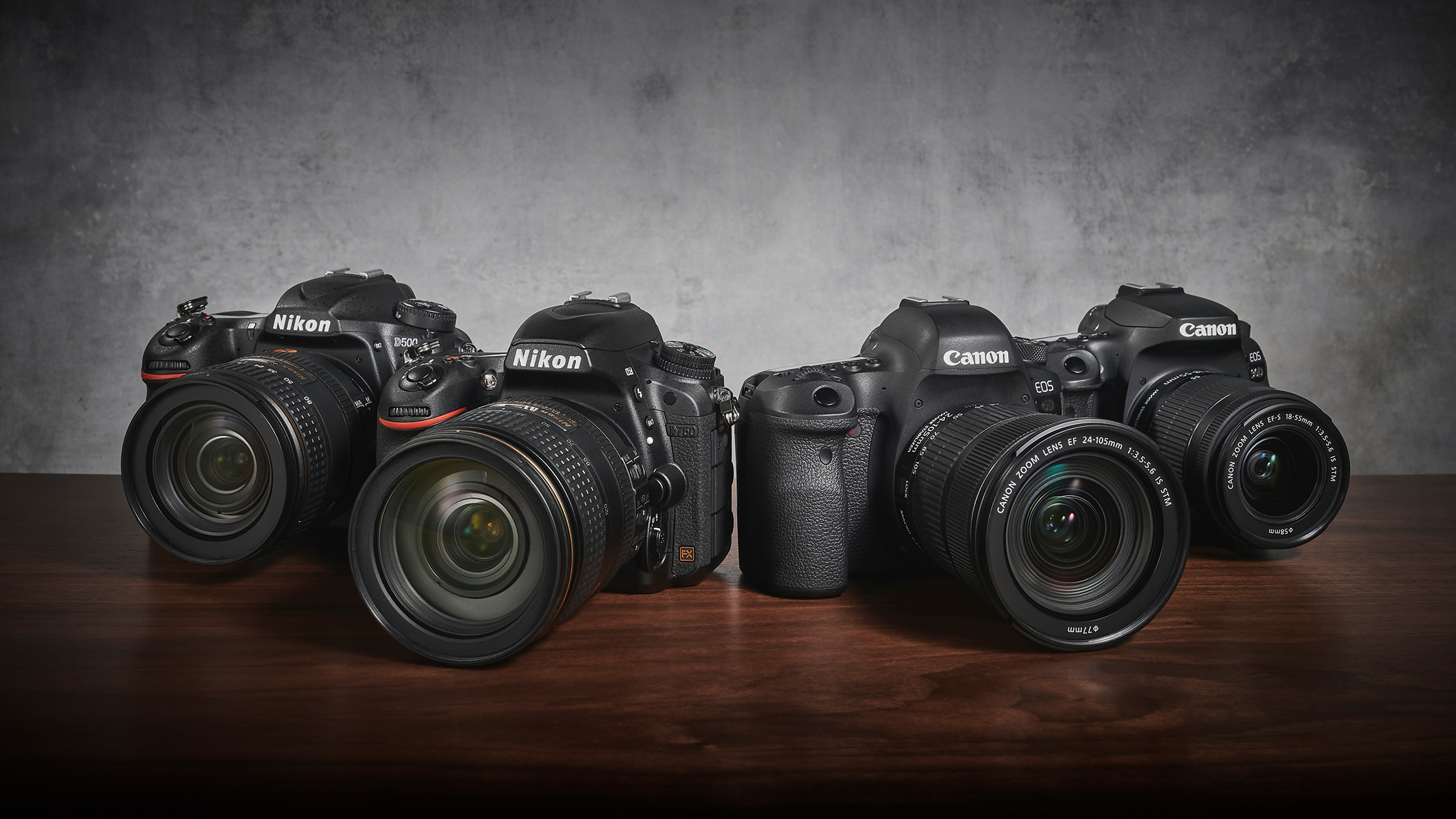 A side-by-side display of two Nikon cameras and two Canon cameras on a wooden table, showcasing their lenses against a textured background