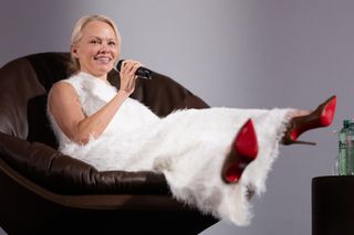 Pamela Anderson wearing a fuzzy white top and pants with red heels sitting in a circle chair and talking into a mic