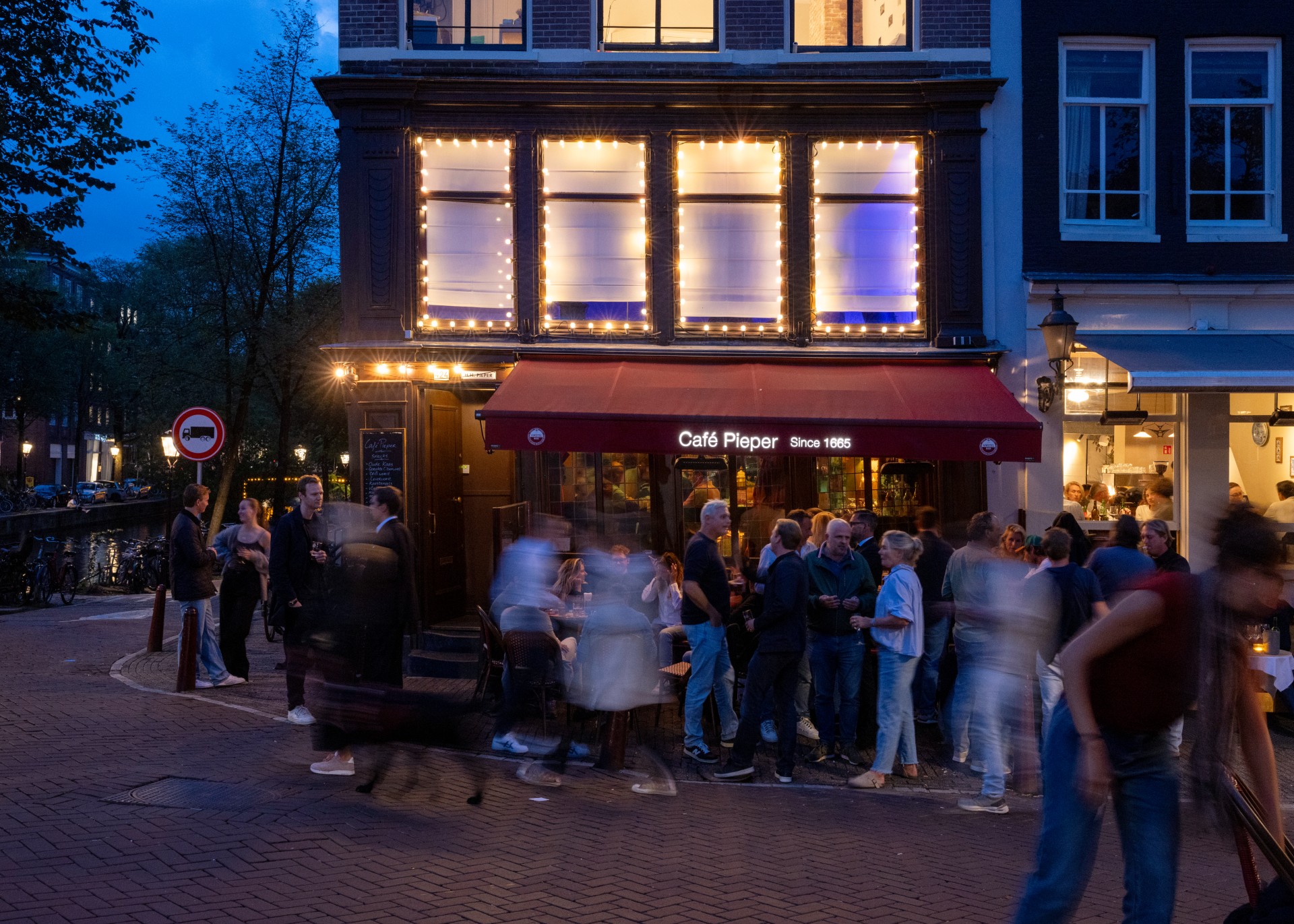 t blue hour, a crowd of drinkers gathers outside Caf&amp;eacute; Pieper in Amsterdam, while passers-by blur into ghostly streaks in the foreground.