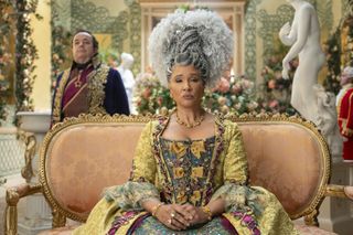 Queen Charlotte (Golda Rosheuvel, center) sits on a couch in an ornate chamber, with Brimsley (Hugh Sachs, left) behind her, in 'Bridgerton' season 4.